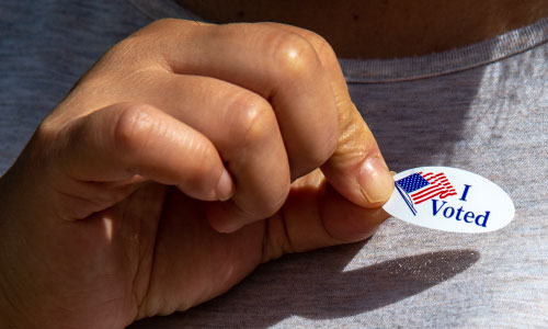 Hand holding an I Voted sticker about to be placed on a grey t-shirt.
